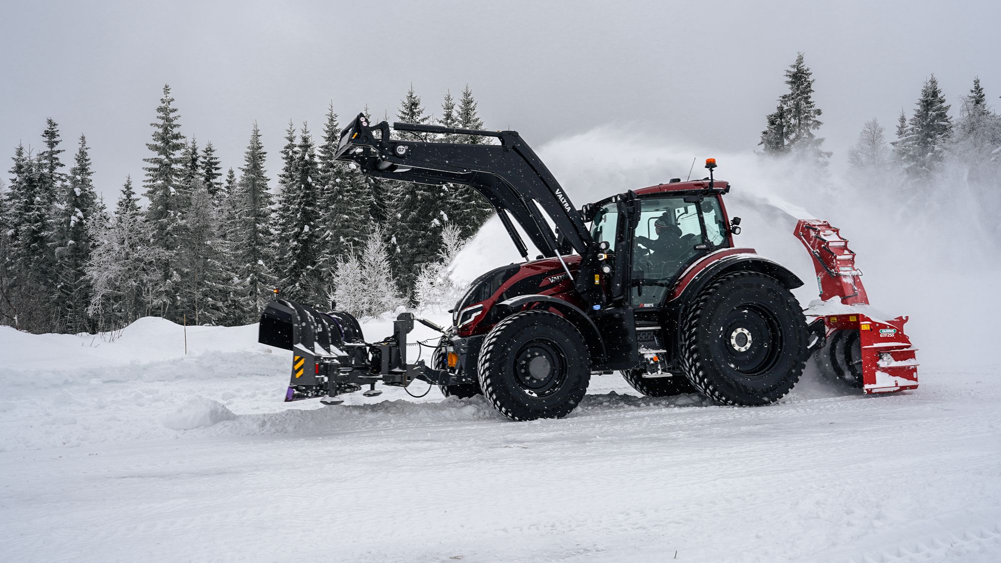 Snøplog montert på traktor rydder snø på gårdsplass i vinterlandskap.